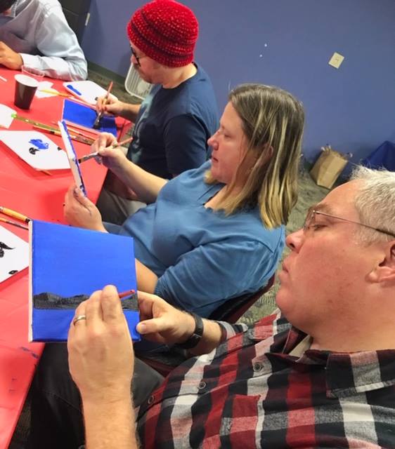 Several people are seated at a table, focused on painting on small canvases. They are holding paintbrushes and working on their artwork, with various shades of blue paint visible on their canvases. The setting appears to be an indoor art class or workshop.