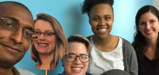 Four smiling people and a cardboard cutout against a light blue background