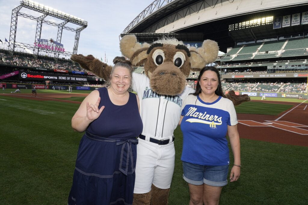 Two women are standing on a baseball field with the Mariners' mascot, a moose, in the center. The woman on the left is making a sign language gesture and smiling, while the woman on the right is also smiling. The stadium is visible in the background.