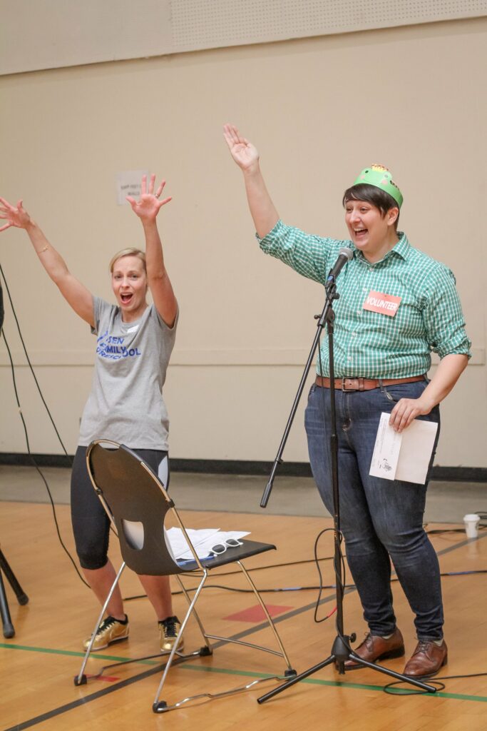 Two people are standing in a room, both raising their arms and smiling. One person on the left is wearing a gray t-shirt and shorts, and the other on the right is wearing a green checkered shirt, a name tag that says "Volunteer," and a green hat. A microphone stand is in front of the person on the right, and a chair with some papers is in the foreground