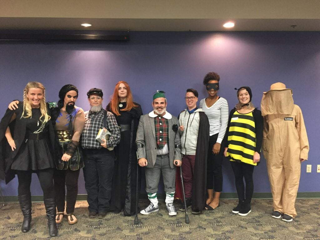 Six people are standing in a row, dressed in various costumes. From left to right: a person dressed as a bearded character in plaid, a person with long red hair in a black outfit, a person in a Scottish-themed costume, a person with short hair and glasses in casual attire, a person in a mask and cape, and a person dressed as a bee. They are indoors against a purple wall.