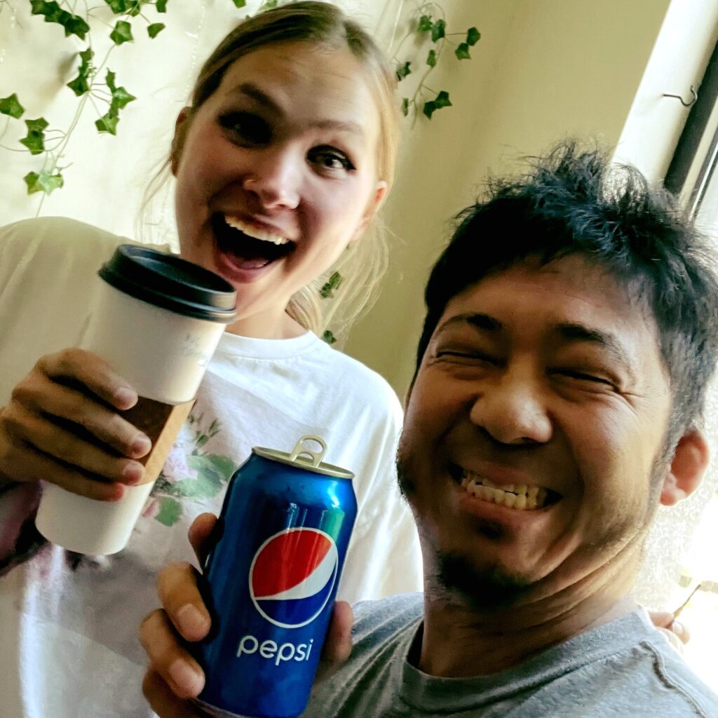 Two people are smiling and holding drinks. The person on the left is holding a coffee cup, while the person on the right is holding a can of Pepsi. They appear to be enjoying themselves in a casual setting with some greenery in the background.