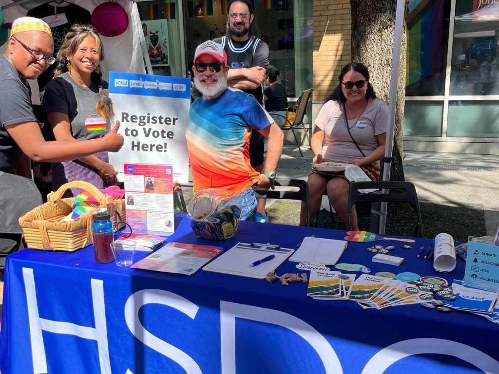 A man wearing a colorful striped shirt and a white cap is standing behind a table with various informational materials. A sign next to him reads "Register to Vote Here!" Two people are sitting behind the table, smiling. The setting appears to be an outdoor event or fair.