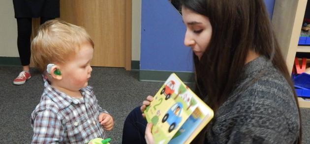 An adult is reading a book to a young child wearing a hearing aid.