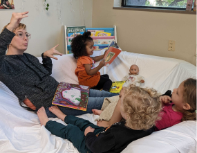 A group of children sits on a couch reading books, with one adult interacting animatedly with them.