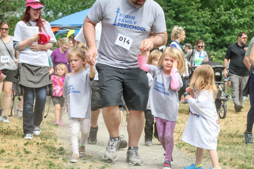 A man wearing a "Rosen Family Preschool" t-shirt and holding the hands of two young girls, who are also wearing matching shirts, is walking in an outdoor event. Other children and adults are visible in the background, participating in the same event.