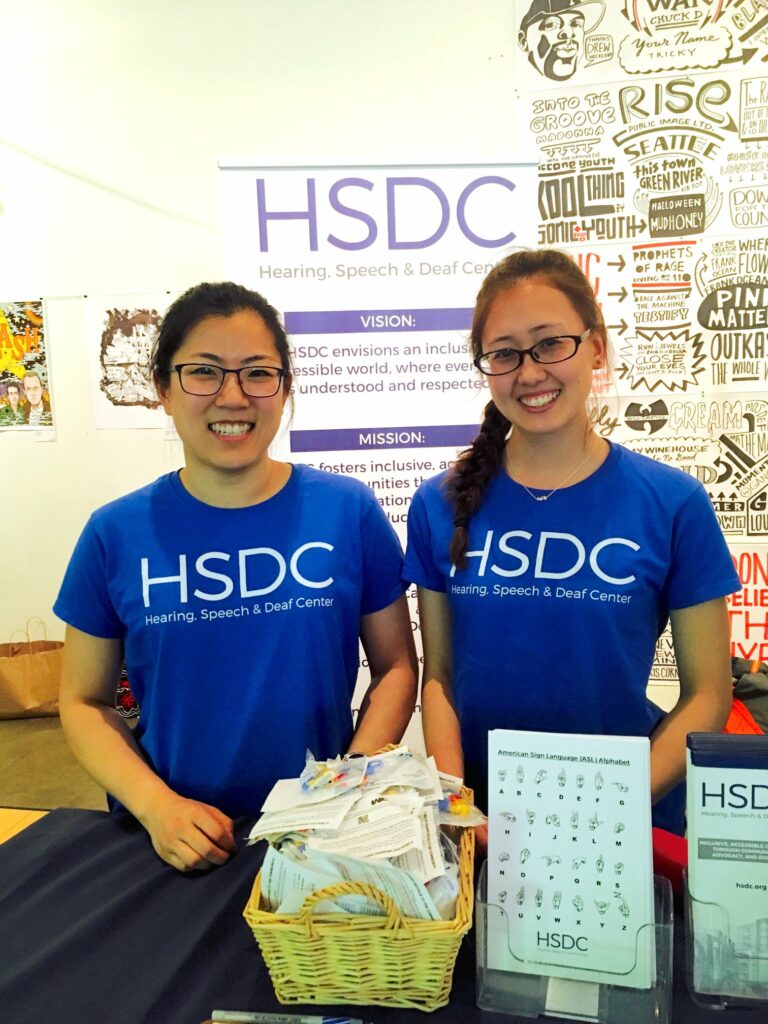 Two women wearing blue "HSDC" t-shirts are standing behind a table with a basket of materials. A sign behind them reads "HSDC Hearing, Speech & Deaf Center" with mission statements. They are smiling and appear to be at an event or information booth.
