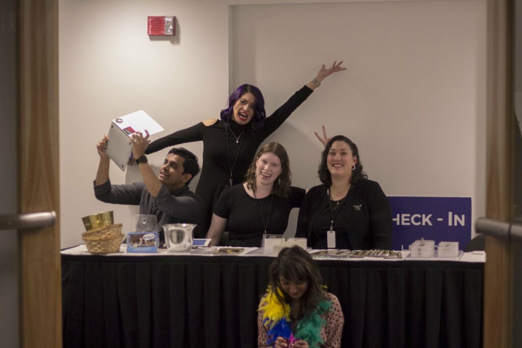 Four people are posing behind a table, smiling and making playful gestures. A woman with colorful feathers is sitting on the floor in front, looking down